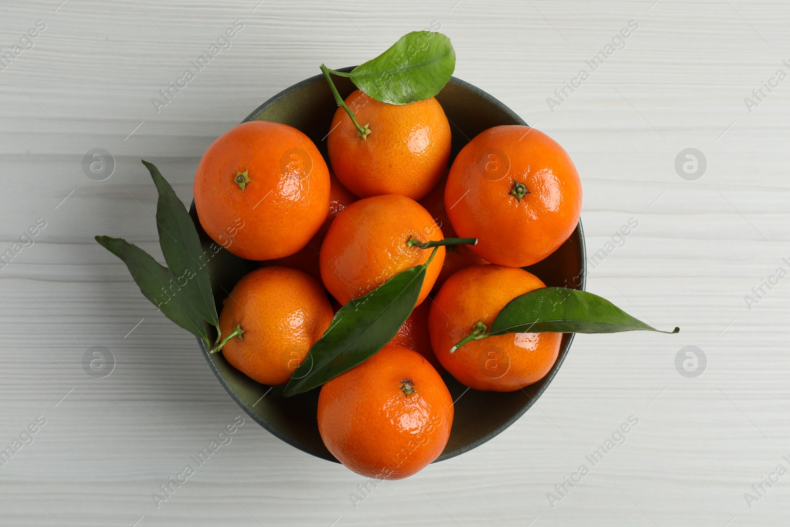 Photo of Delicious tangerines with green leaves in bowl on white wooden table, top view