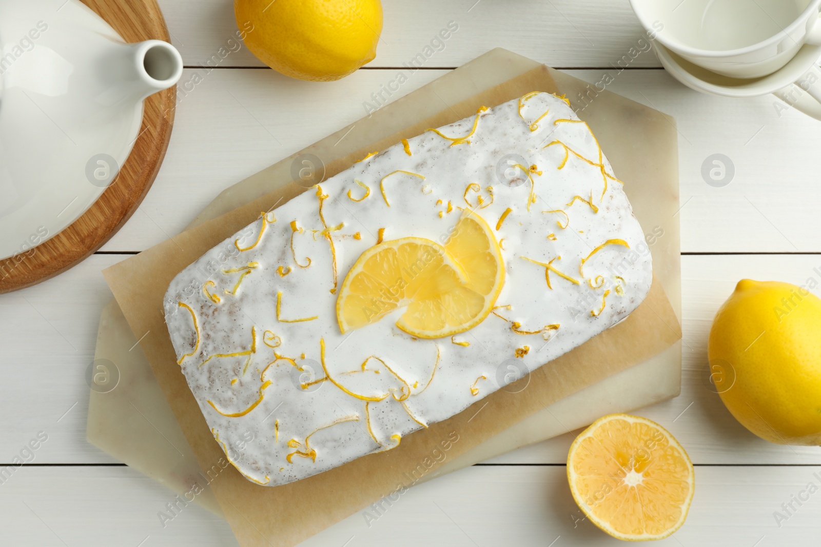 Photo of Tasty lemon cake with glaze, citrus fruits, teapot and cups on white wooden table, flat lay