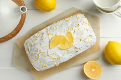 Tasty lemon cake with glaze, citrus fruits, teapot and cups on white wooden table, flat lay