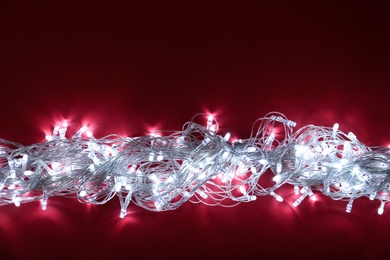 Photo of Glowing Christmas lights on burgundy background, top view. Space for text