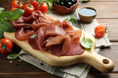 Board with delicious bresaola served with tomato and basil leaves on wooden table, closeup