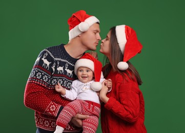 Happy couple with cute baby in Christmas outfits and Santa hats on green background