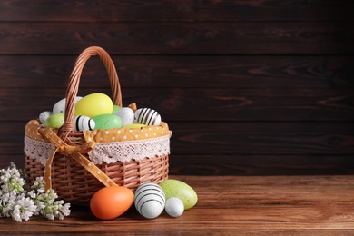 Wicker basket with festively decorated Easter eggs and white lilac flowers on wooden table. Space for text
