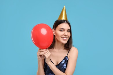 Happy young woman in party hat with balloon on light blue background