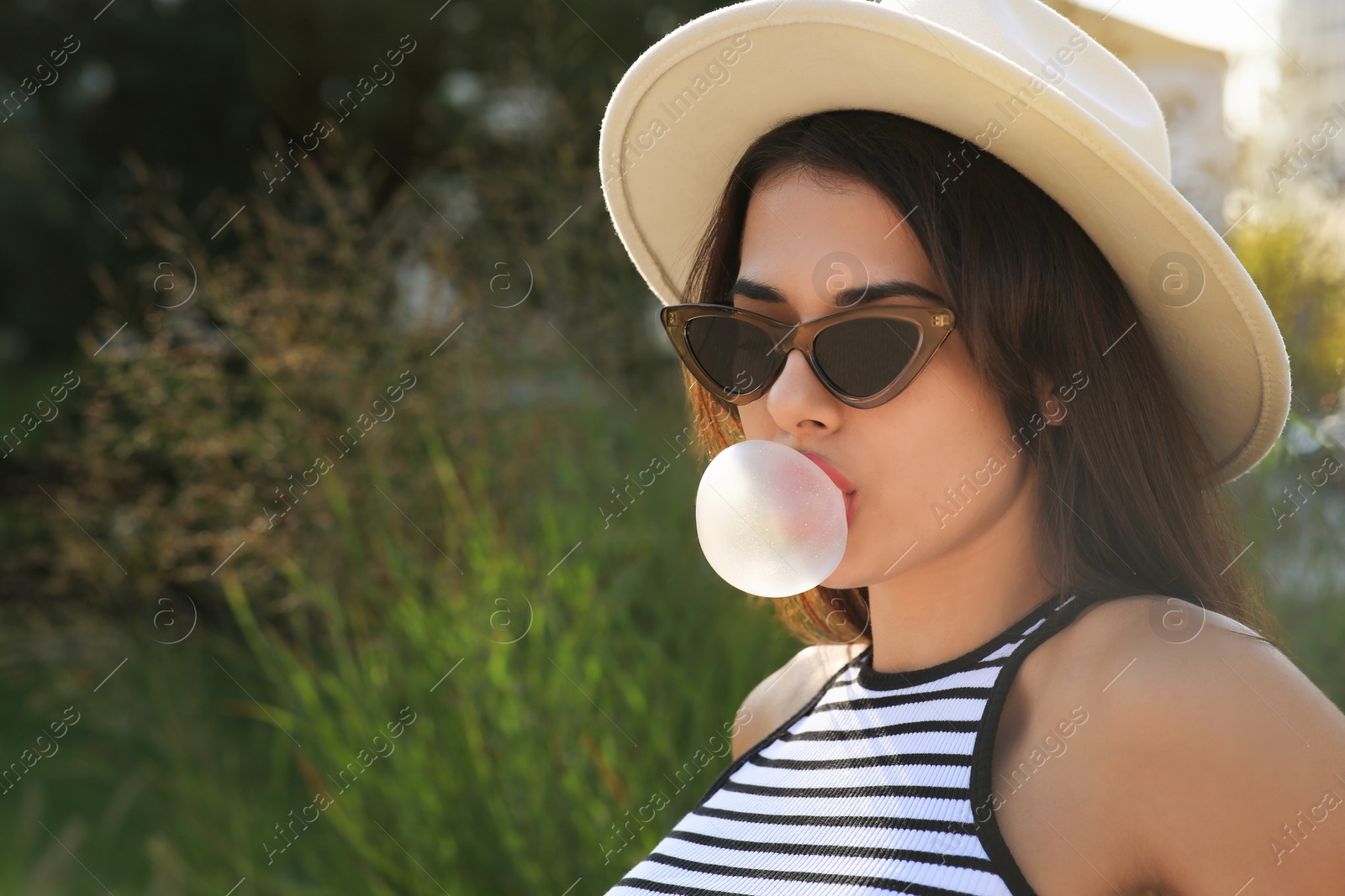 Photo of Beautiful woman in stylish sunglasses blowing gum outdoors on sunny day, space for text