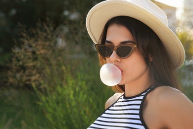 Beautiful woman in stylish sunglasses blowing gum outdoors on sunny day, space for text