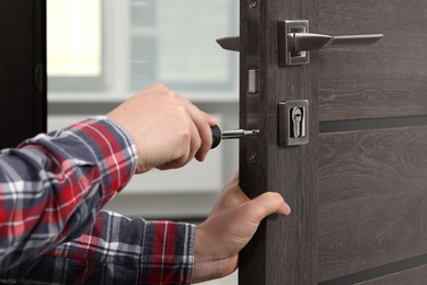 Photo of Handyman with screwdriver repairing door lock indoors, closeup