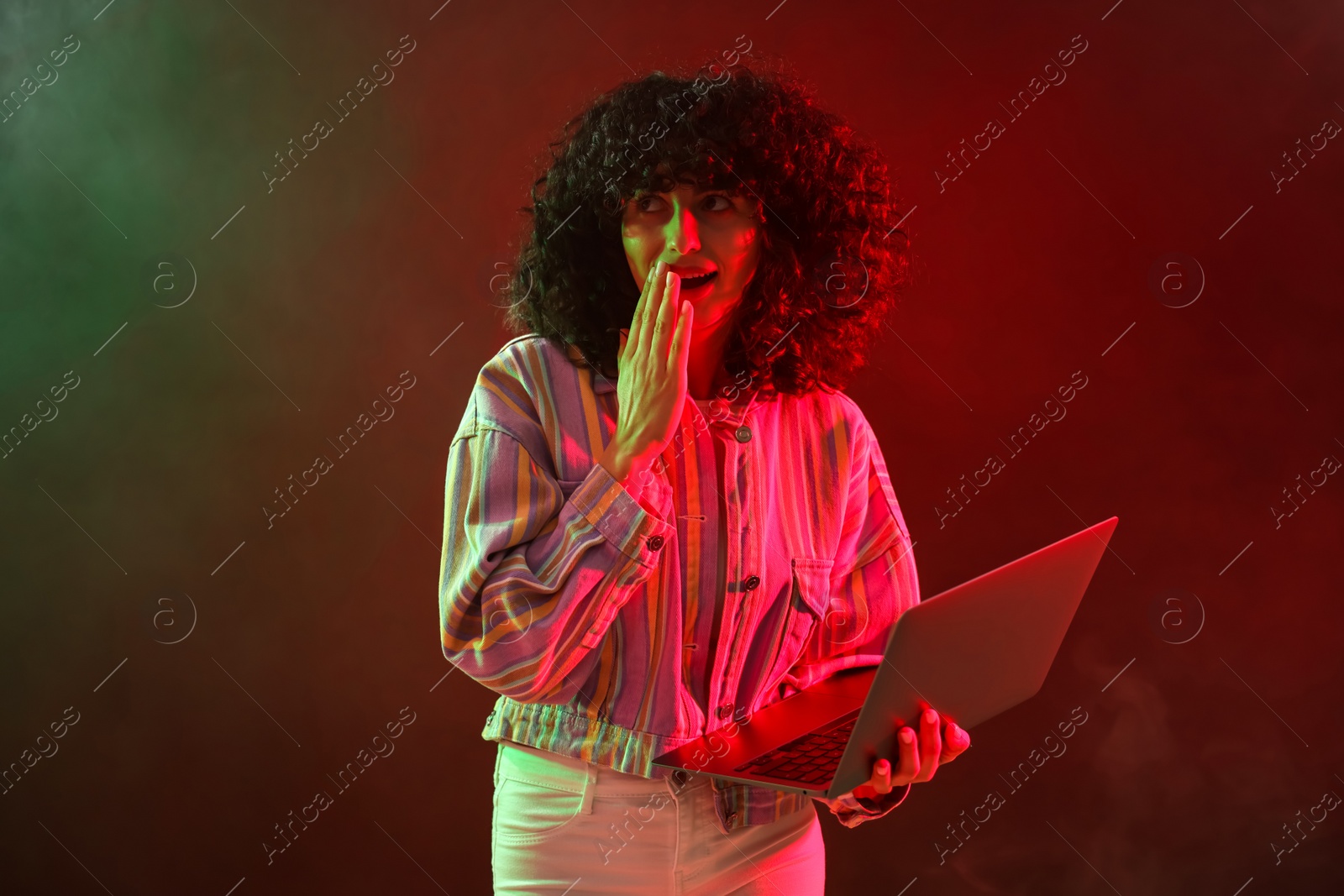 Photo of Beautiful young woman with laptop on color background in neon lights and smoke