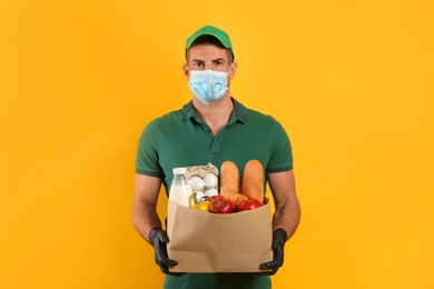 Photo of Courier in medical mask holding paper bag with food on yellow background. Delivery service during quarantine due to Covid-19 outbreak