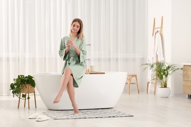 Photo of Beautiful happy woman in stylish robe with cup of hot drink sitting on tub in bathroom