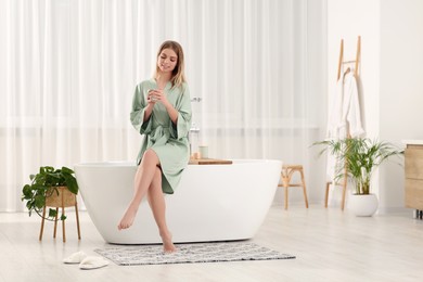 Beautiful happy woman in stylish robe with cup of hot drink sitting on tub in bathroom