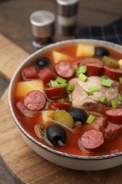 Photo of Meat solyanka soup with thin dry smoked sausages in bowl on wooden board, closeup