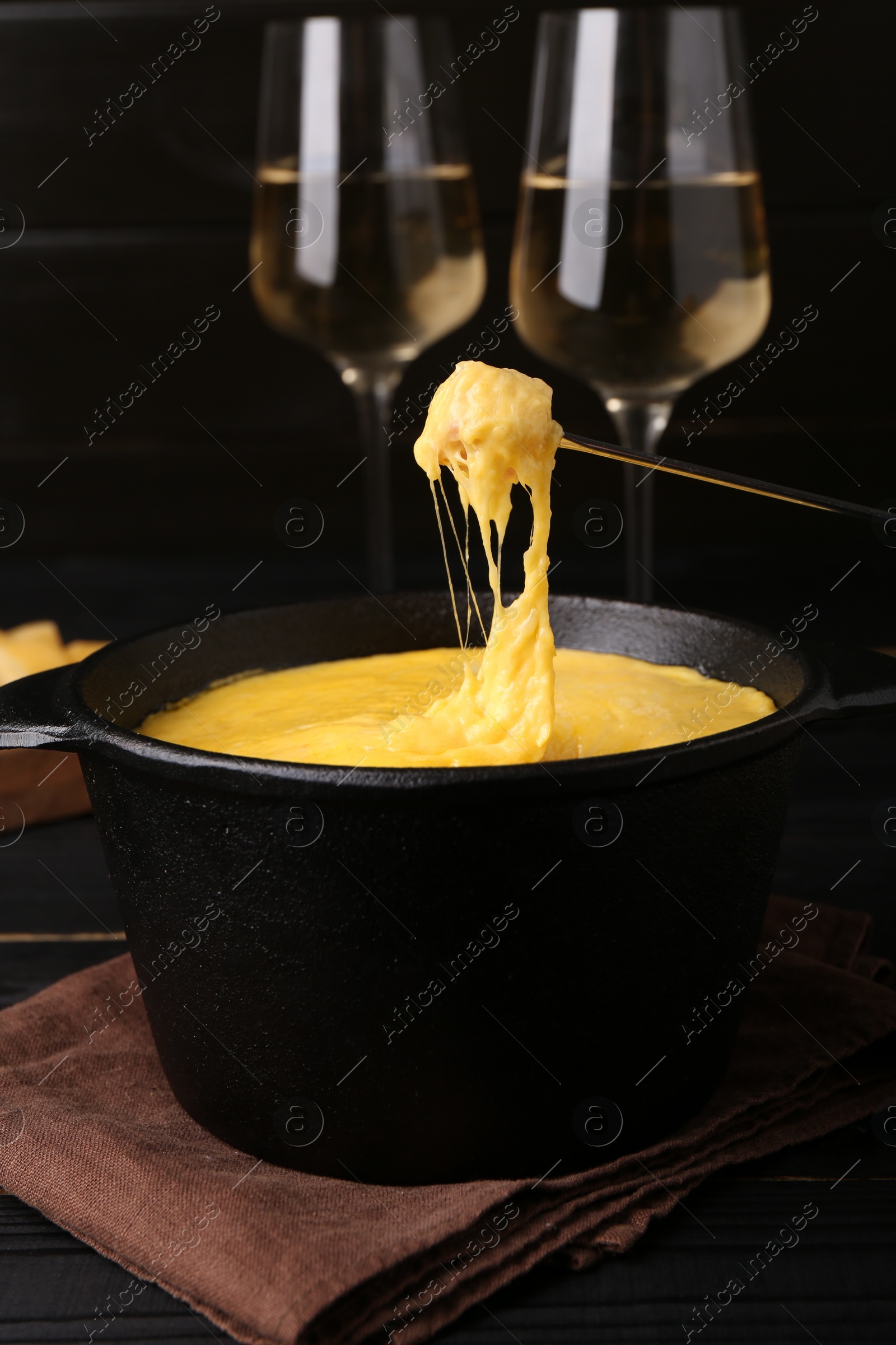 Photo of Dipping piece of ham into fondue pot with melted cheese on black wooden table