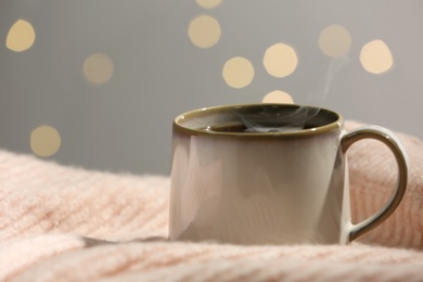 Cup of hot drink on pink sweater against blurred lights, closeup. Cozy atmosphere