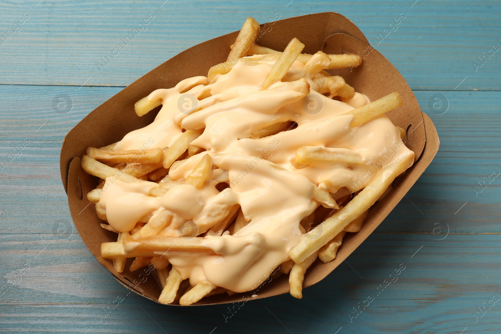 Photo of Tasty potato fries and cheese sauce in paper container on light blue wooden table, top view