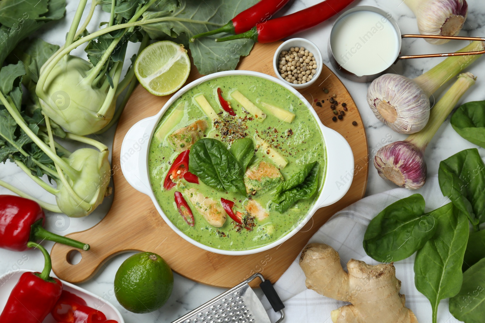 Photo of Saucepan with delicious green curry chicken soup and different ingredients on white marble table, flat lay