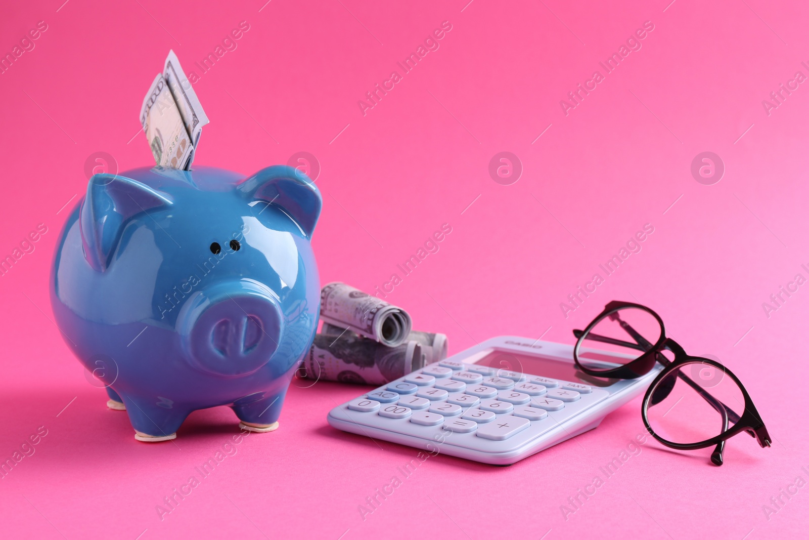 Photo of Financial savings. Piggy bank, dollar banknotes, glasses and calculator on pink background