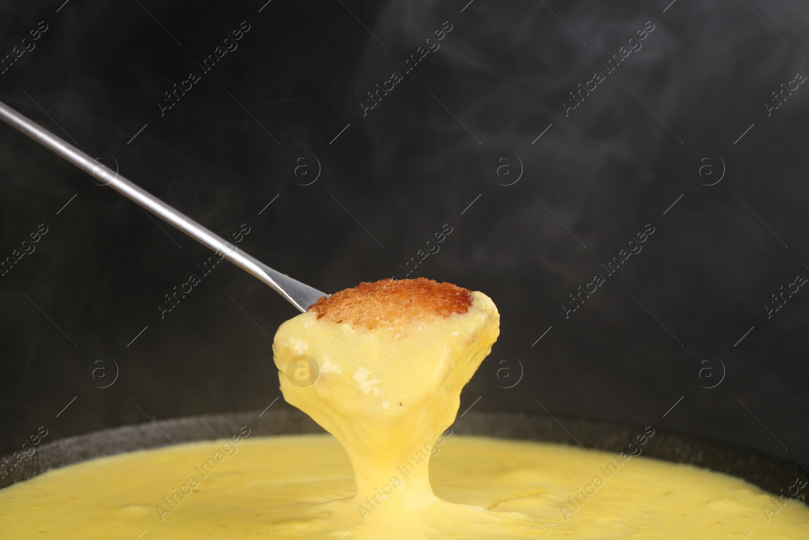 Photo of Dipping piece of bread into fondue pot with tasty melted cheese against dark gray background, closeup