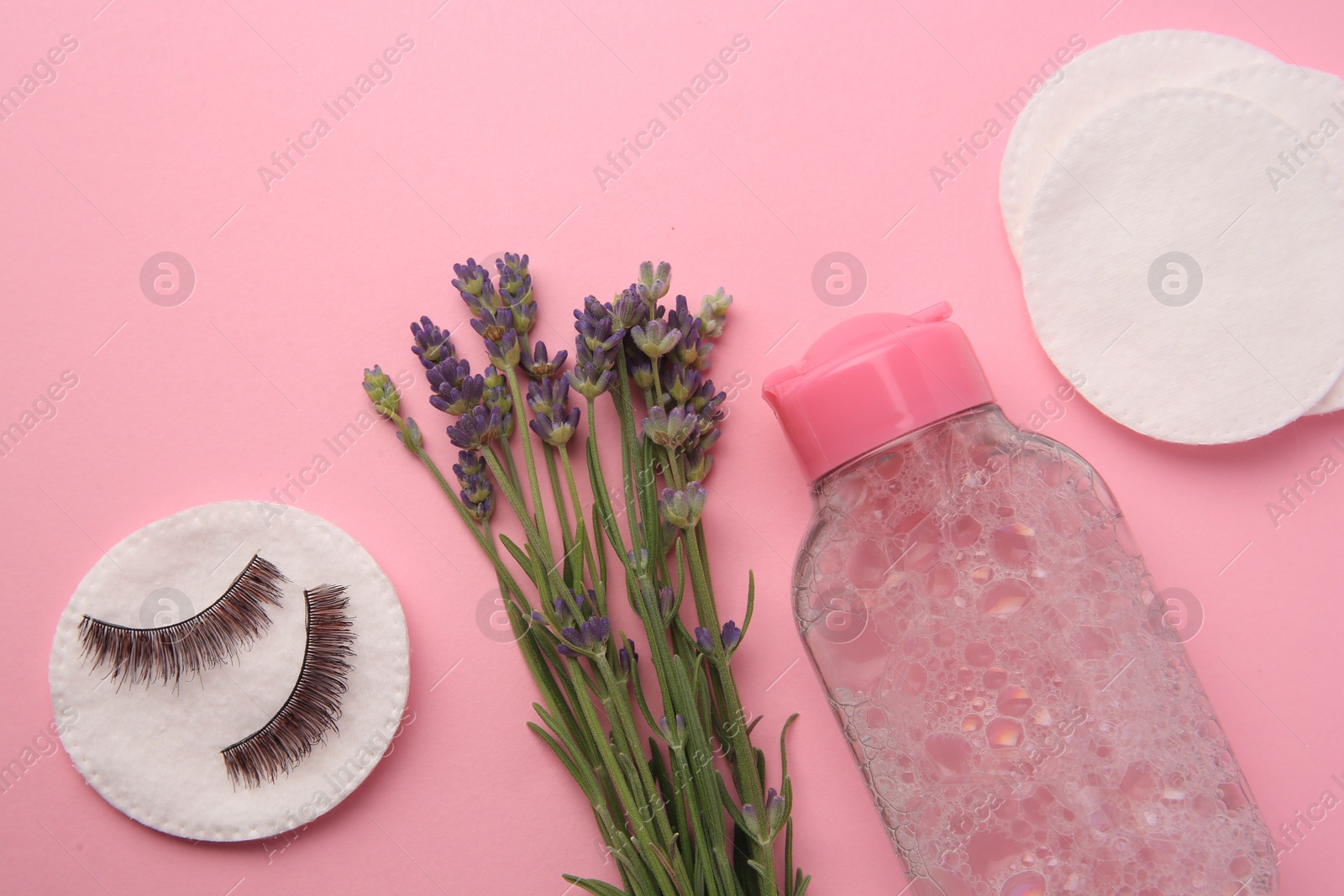 Photo of Bottle of makeup remover, lavender, cotton pads and false eyelashes on pink background, flat lay