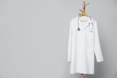 Medical uniform and stethoscope hanging on rack against light grey background. Space for text