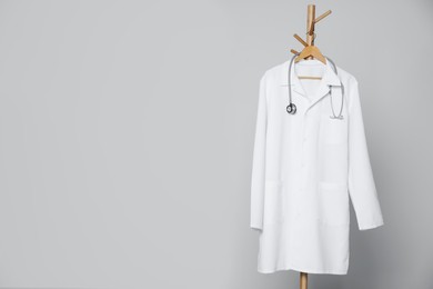 Photo of Medical uniform and stethoscope hanging on rack against light grey background. Space for text