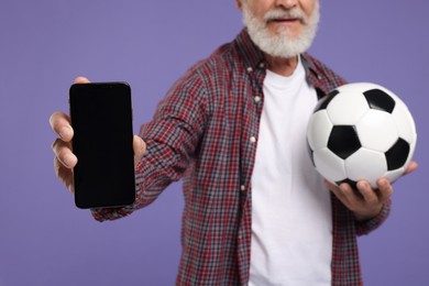 Photo of Sports fan with soccer ball showing smartphone on purple background, closeup