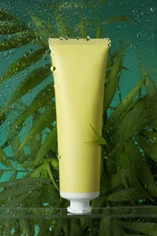 Tube with moisturizing cream and palm leaves on green background, view through wet glass