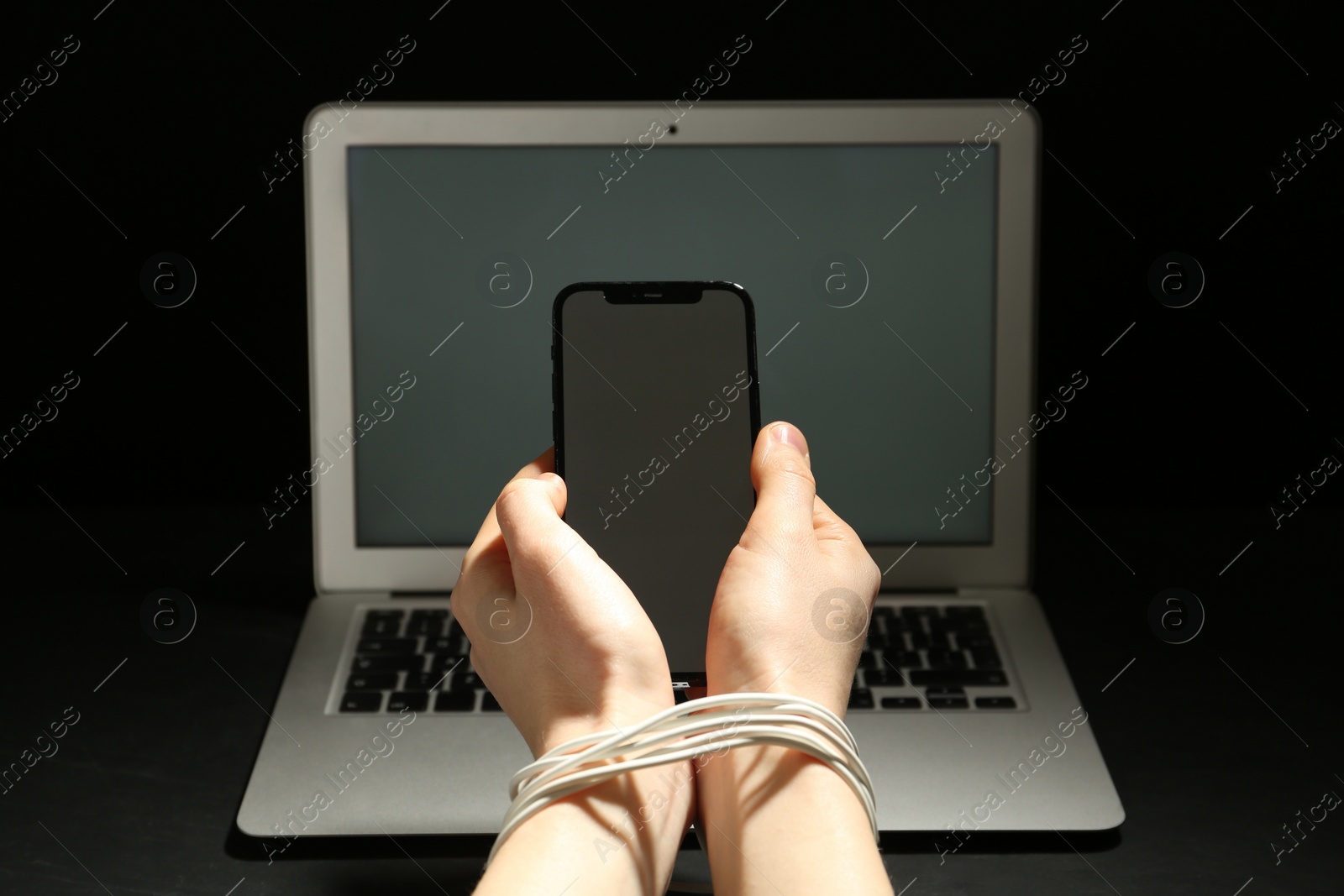 Photo of Woman with tied hands holding smartphone near laptop on dark background, closeup. Internet addiction