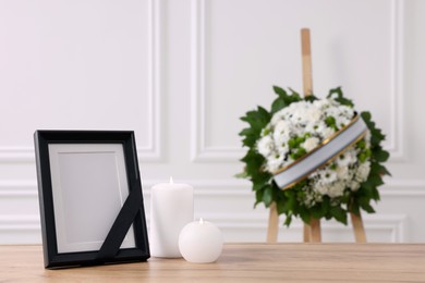 Photo frame with black ribbon, burning candles on table and wreath of flowers near white wall indoors, space for text. Funeral attributes