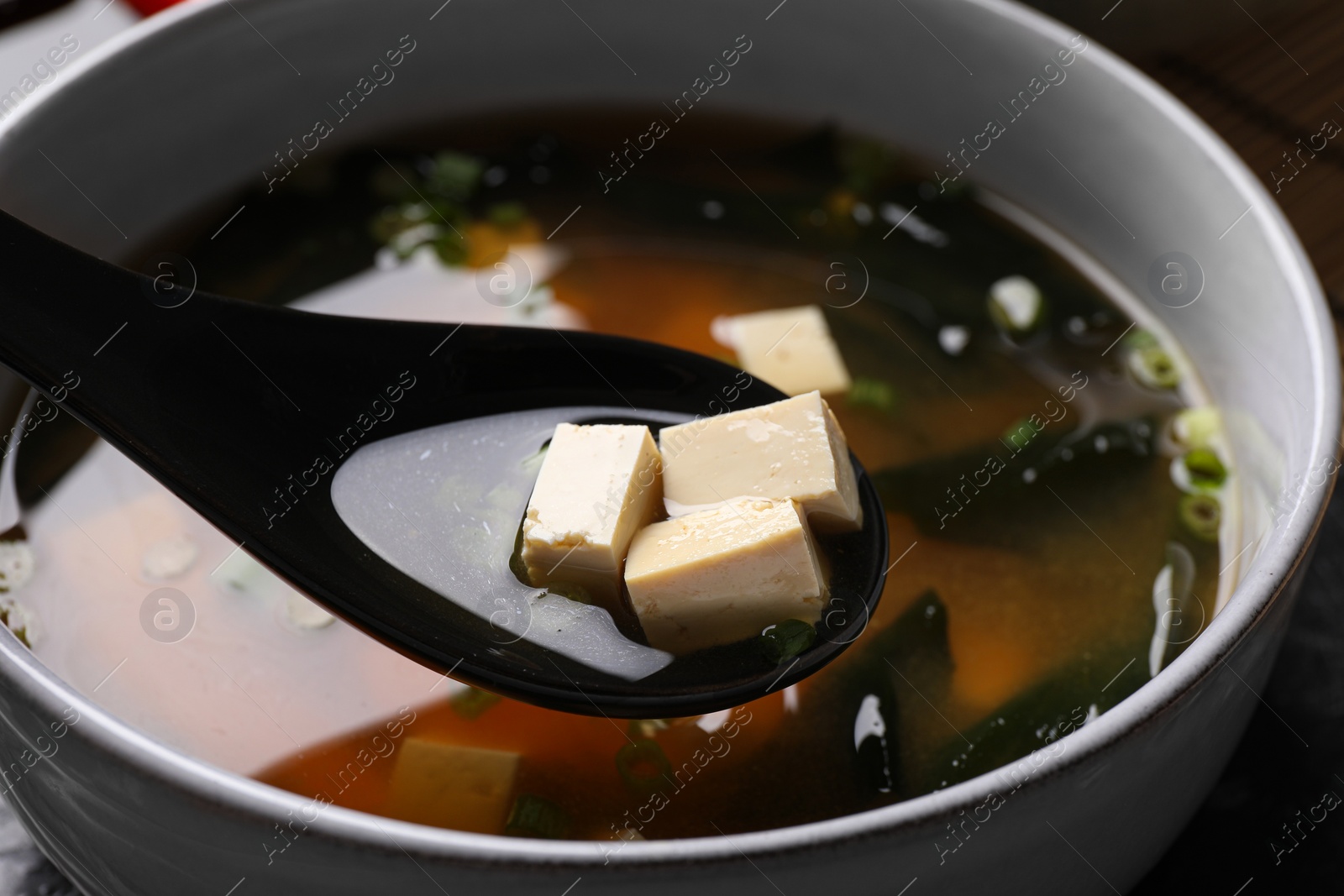 Photo of Delicious miso soup with tofu in spoon above bowl, closeup