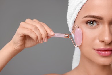 Young woman massaging her face with rose quartz roller on grey background, closeup