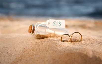 Photo of Gold wedding rings and invitation in glass bottle on sandy beach, closeup