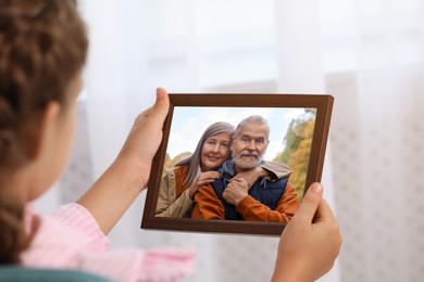 Image of Little girl holding photo frame with family portrait of her grandparents indoors, closeup