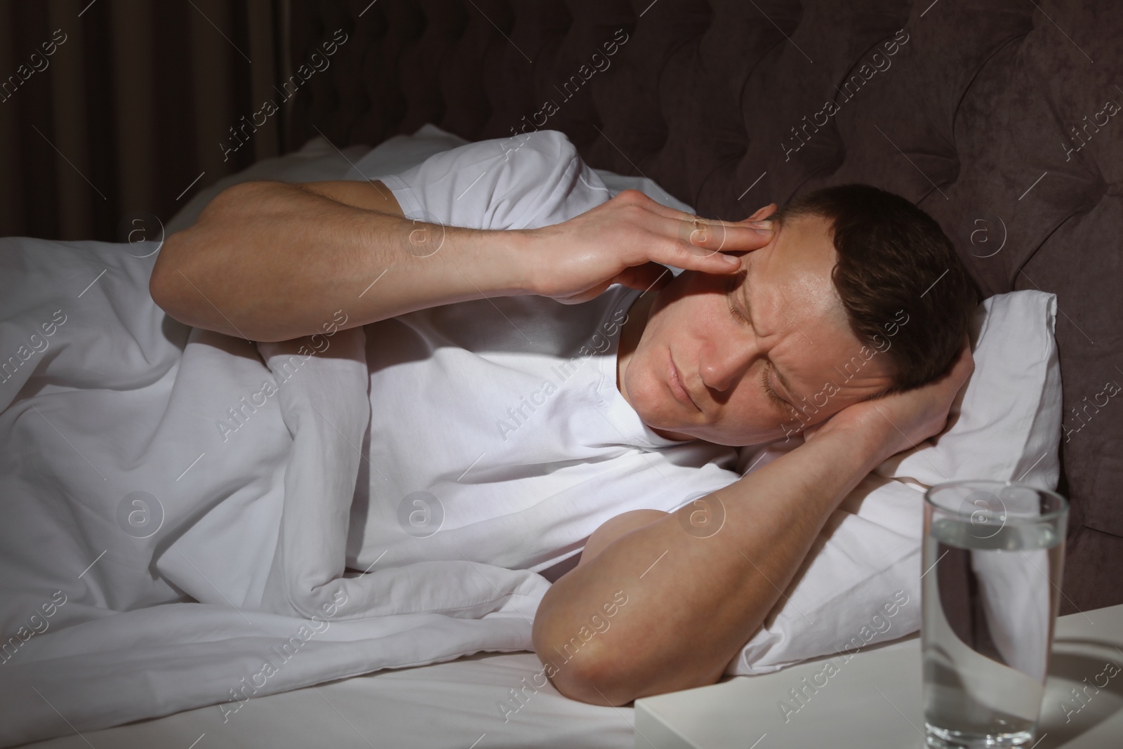 Photo of Handsome young man suffering from headache while lying in bed at night