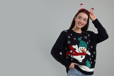 Happy young woman in Christmas sweater and Santa headband on grey background. Space for text