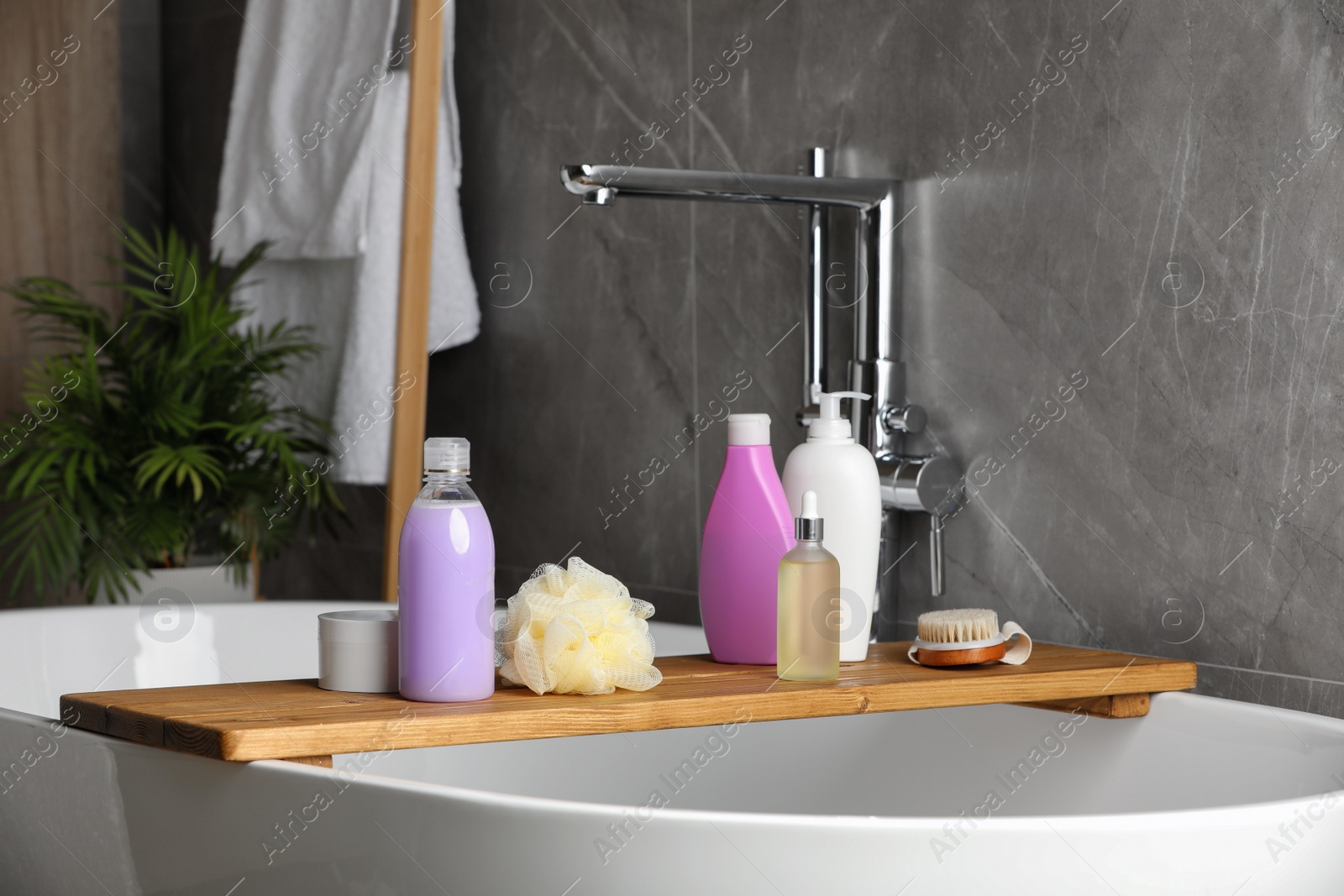 Photo of Wooden bath tray with shampoo, hair oil and other toiletries on tub indoors