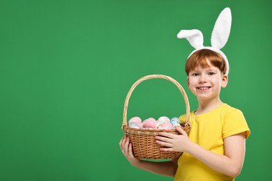 Photo of Easter celebration. Cute little boy with bunny ears and wicker basket full of painted eggs on green background. Space for text