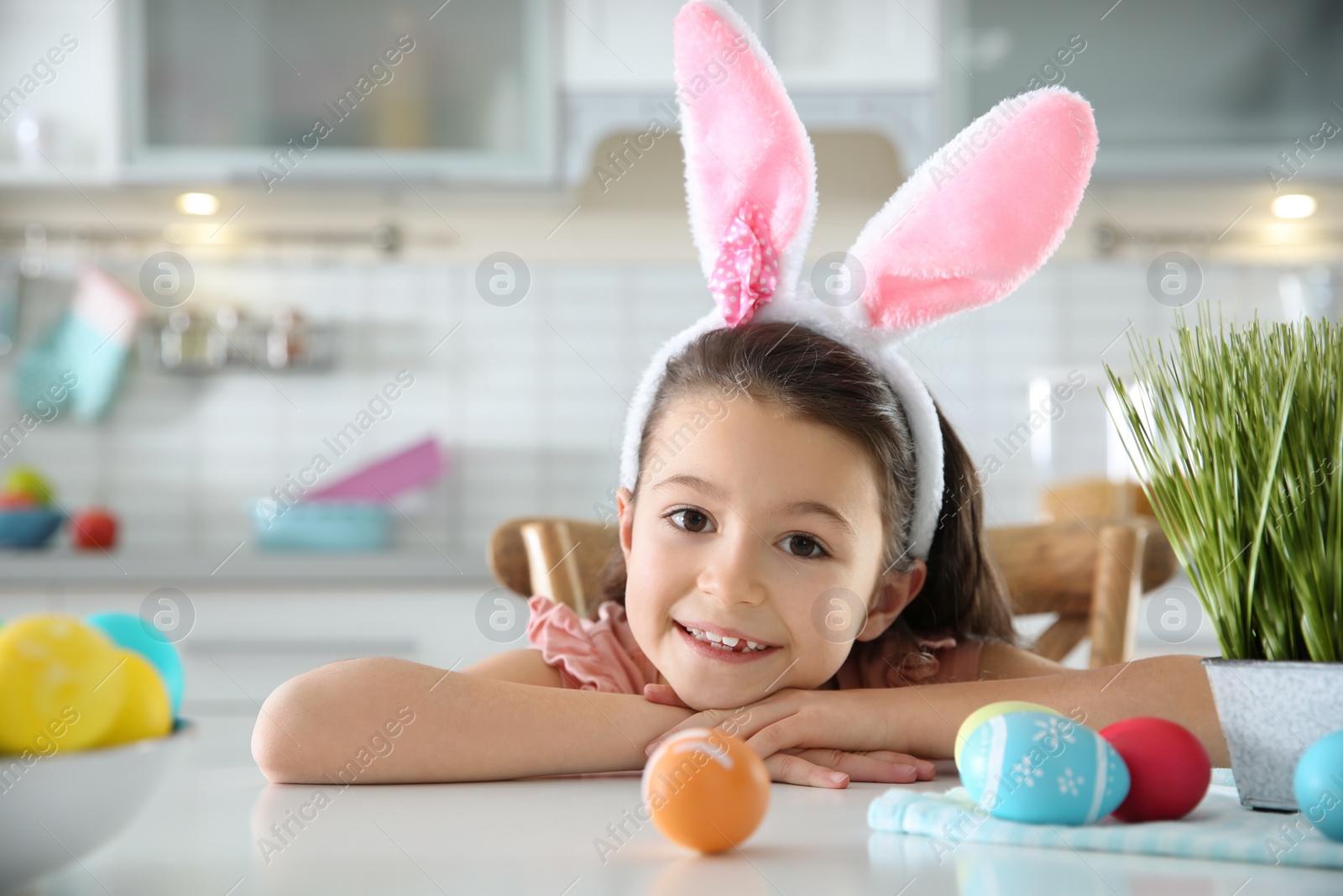 Photo of Cute little girl with bunny ears headband and painted Easter eggs sitting at table in kitchen, space for text