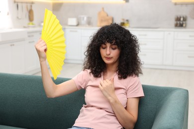 Photo of Young woman waving yellow hand fan to cool herself on sofa at home