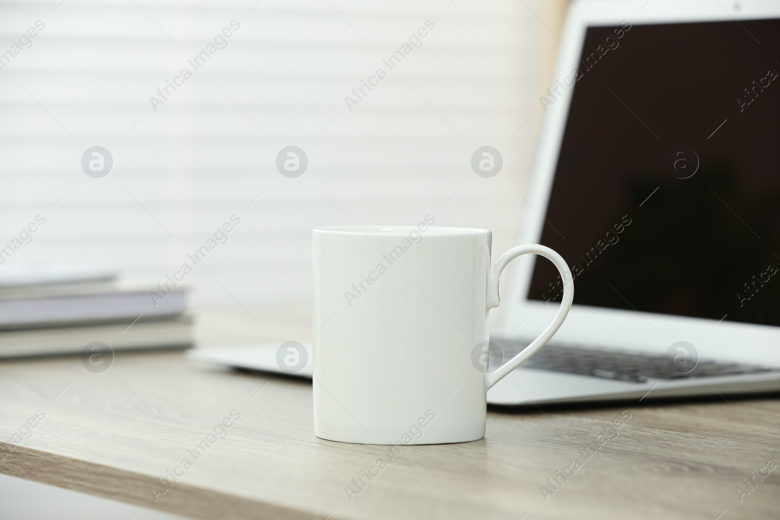 Photo of White ceramic mug and laptop on wooden table indoors