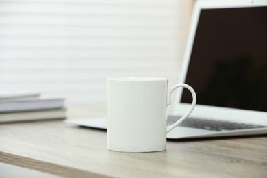 Photo of White ceramic mug and laptop on wooden table indoors