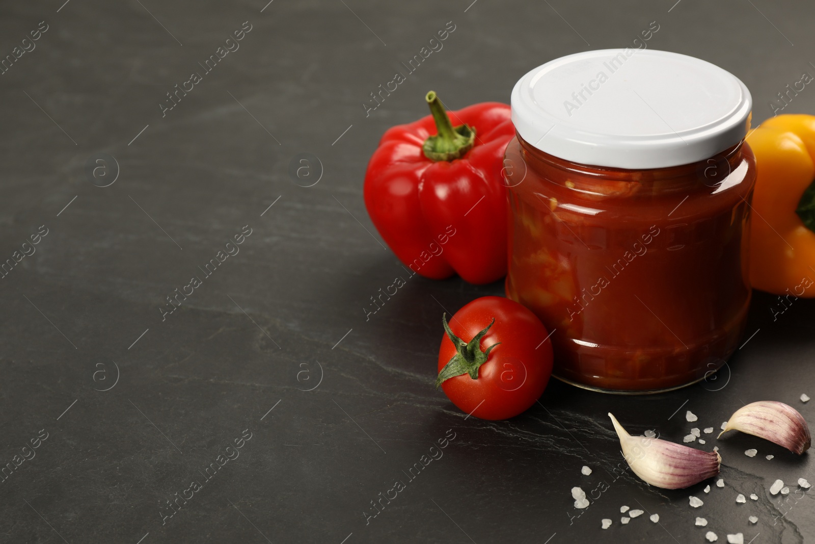 Photo of Glass jar of delicious canned lecho and fresh ingredients on black table. Space for text