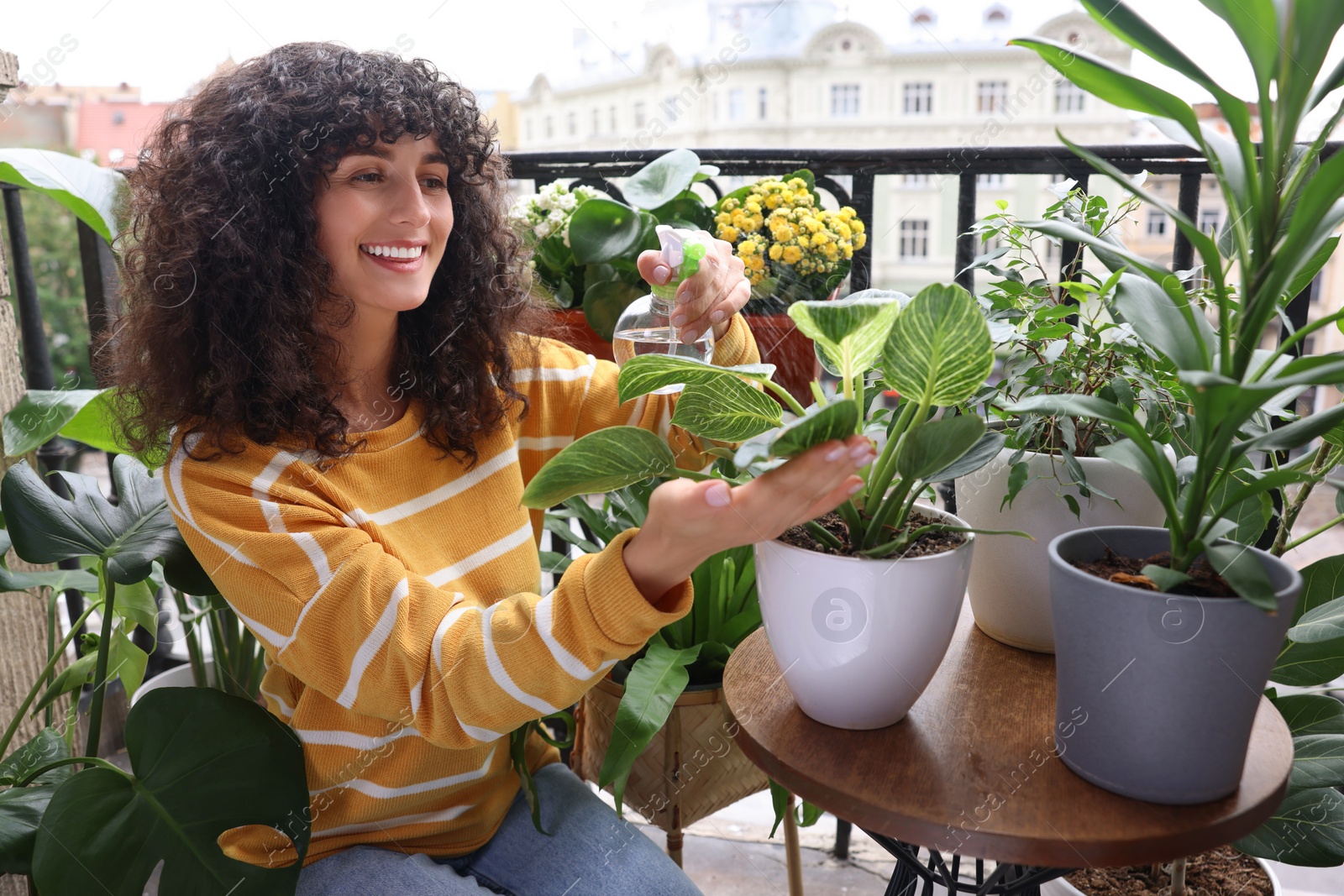 Photo of Beautiful young woman spraying potted houseplants with water on balcony