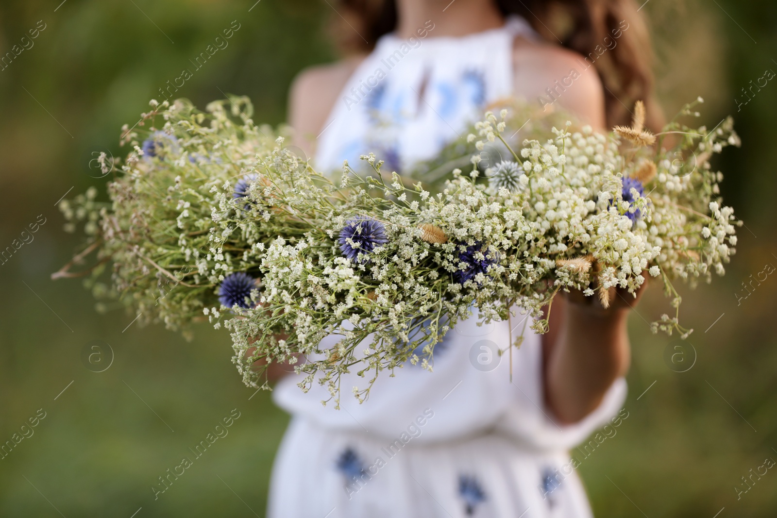 Photo of Little girl holding wreath made of beautiful flowers outdoors, closeup