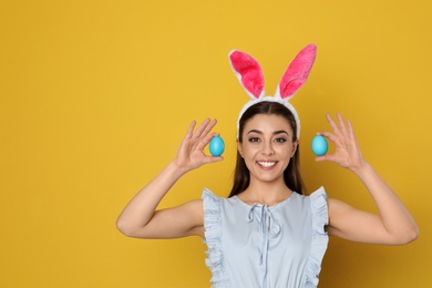 Beautiful woman in bunny ears headband holding Easter eggs on color background, space for text