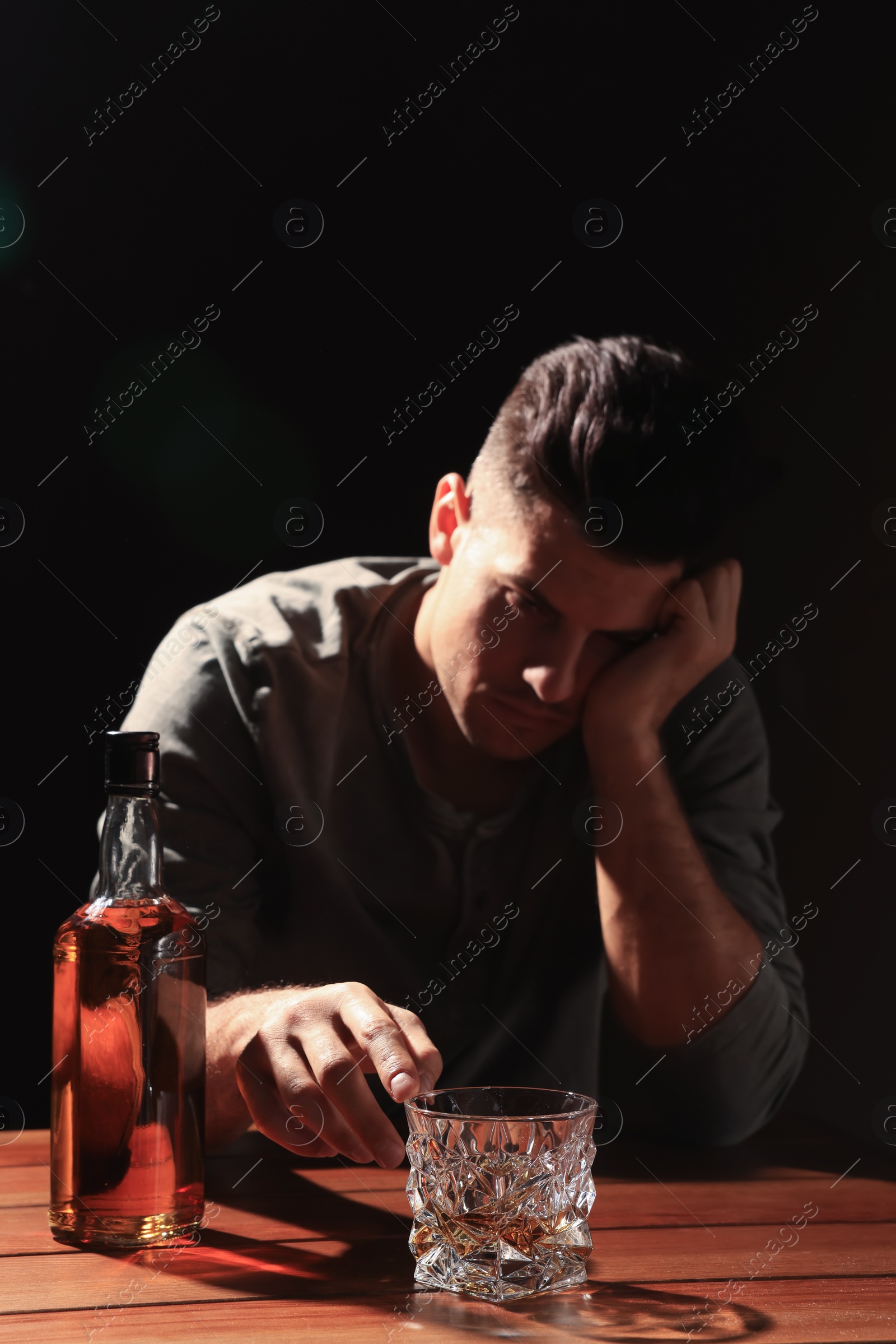 Photo of Addicted man at wooden table against black background, focus on glass of alcoholic drink