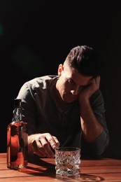 Photo of Addicted man at wooden table against black background, focus on glass of alcoholic drink
