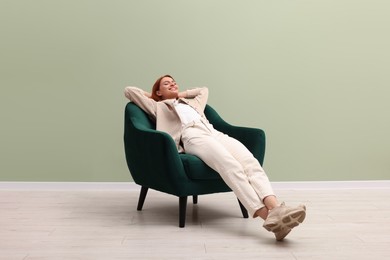 Happy young woman relaxing in armchair near light olive wall indoors