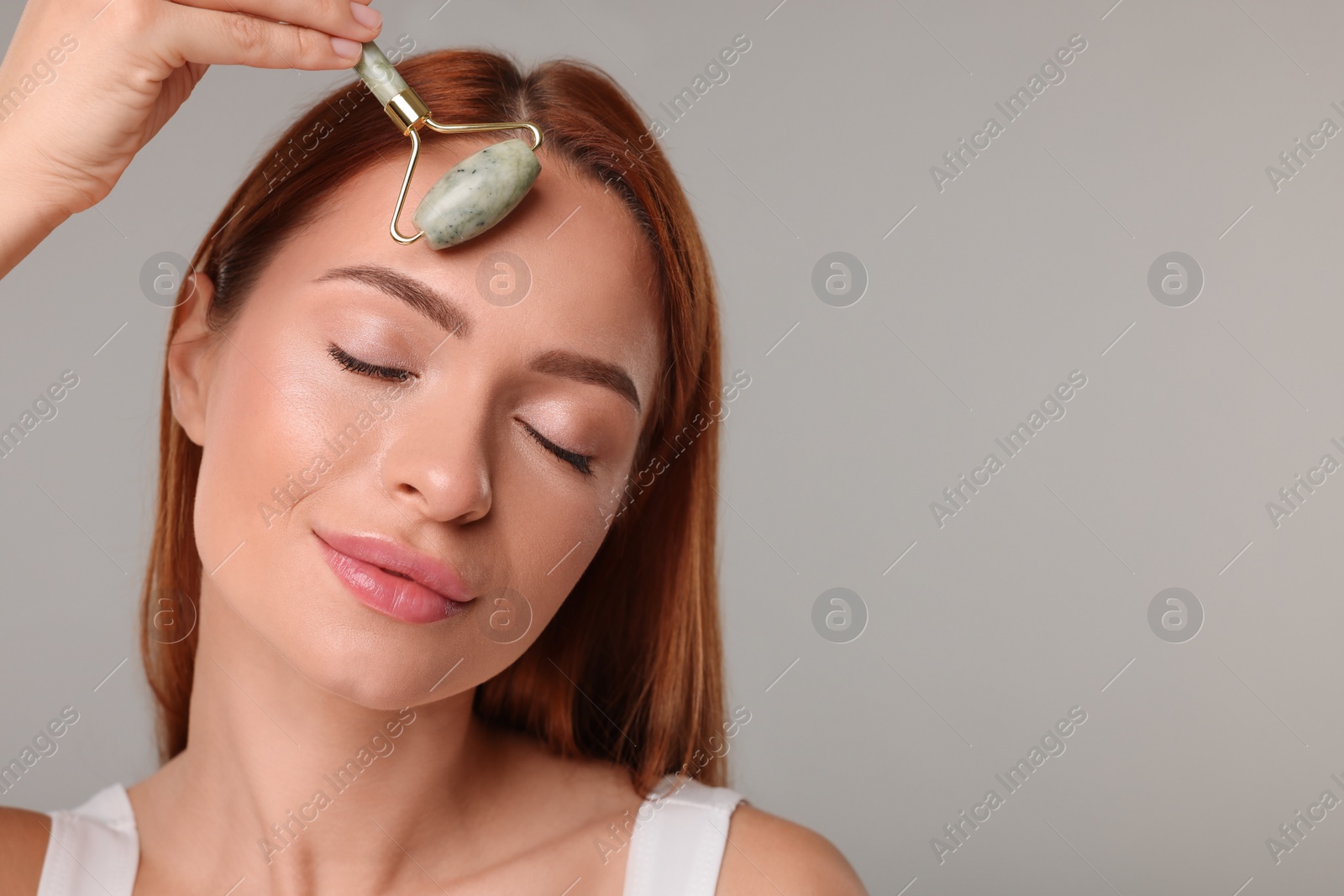 Photo of Young woman massaging her face with jade roller on grey background, space for text