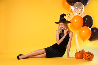 Beautiful woman in witch costume with balloons and pumpkins on yellow background, space for text. Halloween party